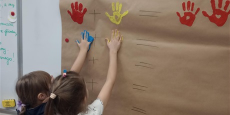 Powiększ grafikę: Uczniowie oddziału przedszkolnego odciskają dłonie zanurzone w farbie na papierze.