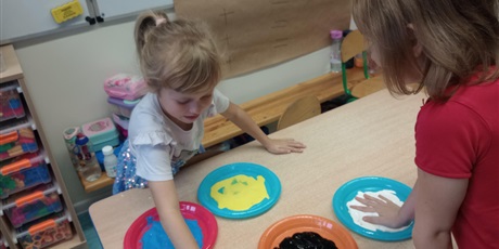 Powiększ grafikę: Dziewczynki zamaczają dłonie w farbach wylanych na talerzyki. Są radosne.