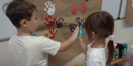 Powiększ grafikę: Uczniowie oddziału przedszkolnego przyciskają do siebie dłonie zanurzone w farbie.