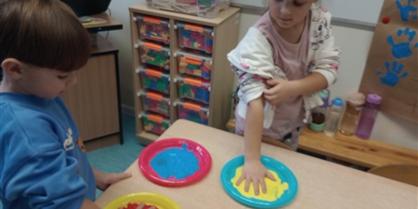 Powiększ grafikę: Dzieci zamaczają dłonie w farbach wylanych na talerzyki. Są radosne.