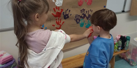 Powiększ grafikę: Uczniowie oddziału przedszkolnego przyciskają do siebie dłonie zanurzone w farbie.