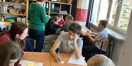 Powiększ grafikę: Uczniowie pracują przy stolikach. Zapisują coś na kartkach.