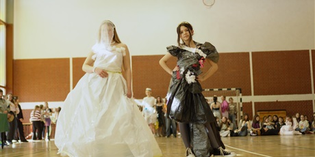 Powiększ grafikę: Zdjęcie ukazuje dwie uczennice prezentujące swoje stroje w pokazie mody ekologicznej. Jedna dziewczynka ubrana jest w białą sukienkę z białym welonem. Obok niej stoi dziewczynka w czarnej sukience.