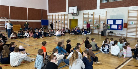 Powiększ grafikę: Zdjęcie przedstawia rozpoczęcie konkursu ekologicznego związanego z obchodami Dnia Ziemi. Uczniowie z klas 4-6 siedzą w grupach. W dali nauczycielka opowiada o historii akcji Dnia Ziemi.