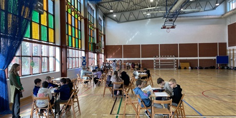 Powiększ grafikę: Na zdjęciu widać uczniów siedzących przy stolikach. Rozwiązują oni test i krzyżówki ekologiczne. Po lewej stronie stoją nauczyciele obserwujący prace uczniów.