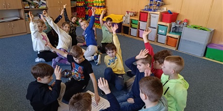 Powiększ grafikę: Grupa dzieci siedzących na dywanie w świetlicy. Dzieci podnoszą ręce do góry.