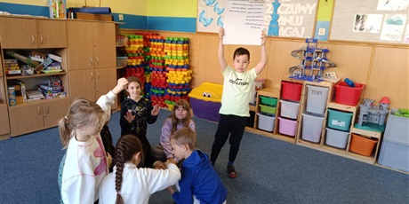 Powiększ grafikę: Grupa dzieci prezentuje efekty swojej pracy. Jeden chłopiec  stoi i trzyma uniesiony do góry plakat.