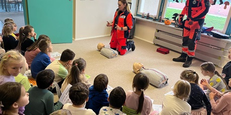 Powiększ grafikę: Pracownik pogotowia pokazuje dzieciom jak udzielać pierwszej pomocy. Klęczy przy manekinie i tłumaczy dzieciom co należy robić.