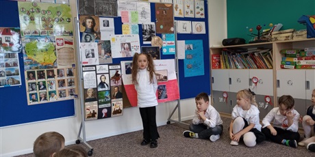 Powiększ grafikę: Dziewczynka stoi przy planszy i opowiada o wybranej osobie. Pozostałe dzieci słuchają.