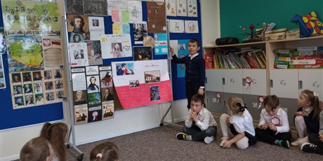 Powiększ grafikę: Chłopiec pokazuje na tablicę. Na niej są zdjęcia znanych Polaków. Opowiada pozostałym dzieciom o jednym z nich.