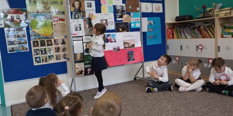 Powiększ grafikę: Dziewczynka wskazuje na swoją pracę. Są na niej informacje o znanym Polaku. Dzieci słuchają opowieści koleżanki.