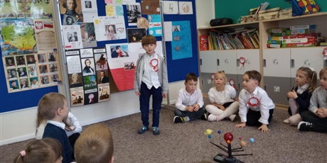 Powiększ grafikę: Chłopiec ubrany w białą bluzkę z przypiętym kotylionem mówi do swoich kolegów. Dzieci siedzą na dywanie i słuchają opowieści.