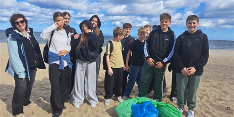 Powiększ grafikę: Uczniowie stoją na plaży. Uśmiechają się. Wcześniej pod okiem nauczycielki stojącej po lewej stronie sprzątali plażę.