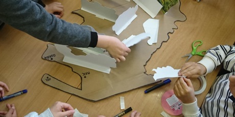 Powiększ grafikę: Na stole leży kontur mapy Kaszub oraz zdjęcia tego regiony. Dzieci układają makietę ze zdjęć.
