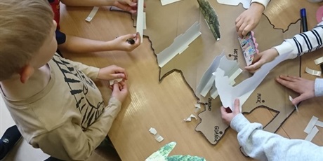 Powiększ grafikę: Na stole leży kontur mapy Kaszub oraz zdjęcia tego regiony. Dzieci układają makietę ze zdjęć.