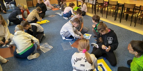 Powiększ grafikę: Dzieci siedzą na dywanie w małych grupach. Układają puzzle z herbami miast kociewskich i przyklejają je na kolorowe kartki.