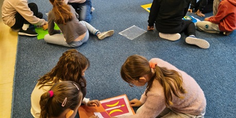 Powiększ grafikę: Dzieci siedzą na dywanie w małych grupach. Układają puzzle z herbami miast kociewskich i przyklejają je na kolorowe kartki.