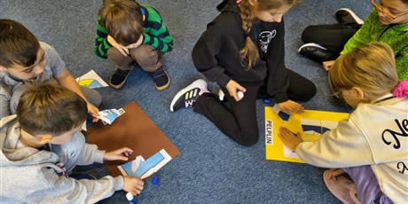 Powiększ grafikę: Dzieci siedzą na dywanie. Układają puzzle z herbami miast kociewskich i przyklejająje na kolorowe kartki.