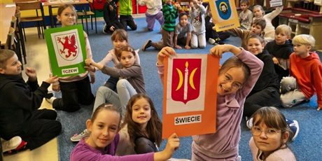 Powiększ grafikę: Dzieci siedzą na dywanie i pozują do zdjęcia. W rękach trzymają kolorowe kartki z herbami miast kociewskich.