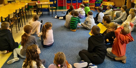 Powiększ grafikę: Dzieci siedzą na dywanie w świetlicy. Zwrócone są twarzą do nauczycielki, która pokazuje region kociewia na ekranie ściennym.