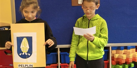 Powiększ grafikę: Dziewczynka i chłopiec stoją na tle granatowej tablicy z napisem KOCIEWIE. Dziewczynka trzyma w rękach kremową kartkę z herbem Pelplina, obok chłopiec trzyma małą karteczkę, z której czyta informację 