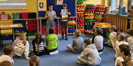 Powiększ grafikę: Dwóch chłopców stoi na tle granatowej tablicy z napisem KOCIEWIE.  Jeden chłopiec trzyma w rękach brązową kartkę z herbem Gniewu, drugi chłopiec trzyma małą karteczkę, z której czyta informację o dany