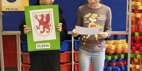 Powiększ grafikę:  Dziewczynka i chłopiec stoją na tle granatowej tablicy z napisem KOCIEWIE. Chłopiec trzyma w rękach zieloną kartkę z herbem Tczewa, obok dziewczynka trzyma małą karteczkę, z której czyta informację o