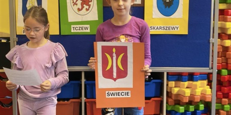 Powiększ grafikę: Dwie dziewczynki stoją na tle granatowej tablicy z napisem KOCIEWIE. Jedna dziewczynka trzyma w rękach pomarańczową kartkę z herbem Świecia, druga dziewczynka trzyma małą karteczkę, z której czyta inf