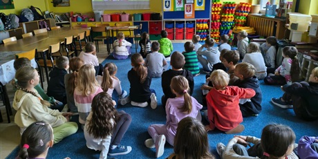 Powiększ grafikę: Duża grupa dzieci siedzi na dywanie tyłem do obiektywu. Dzieci wpatrują się w ekran ścienny.