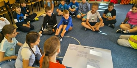 Powiększ grafikę: Grupa dzieci siedzi na dywanie w świetlicy. Na środku leży duży brystol, na którym naklejony jest tekst wiersza o Kociewiu.