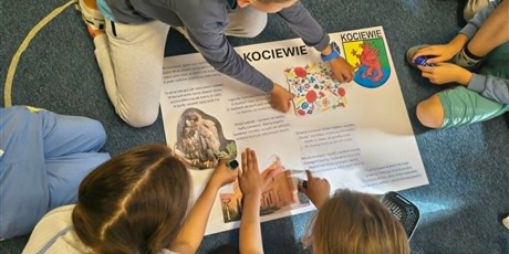 Powiększ grafikę: Grupa dzieci pochyla się nad białym brystolem i przykleja ilustracje związane z Kociewiem.