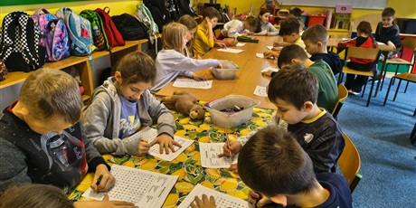 Powiększ grafikę: Grupa dzieci siedzi przy stole w świetlicy. Wszyscy w skupieniu rozwiązują wykreślanki wyrazowe.