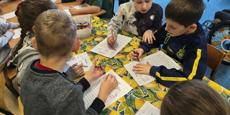 Powiększ grafikę: Grupa dzieci siedzi przy stole w świetlicy. Wszyscy w skupieniu rozwiązują wykreślanki wyrazowe.