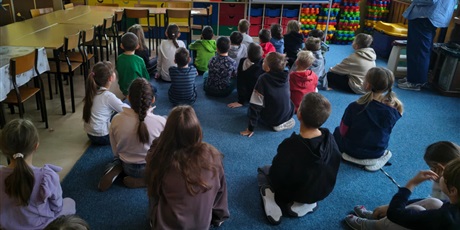 Powiększ grafikę: Grupa dzieci siedzi na dywanie w świetlicy patrząc na film wyświetlany na ekranie ściennym.