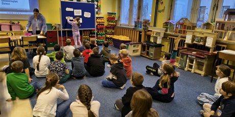 Powiększ grafikę: Grupa dzieci siedzi na dywanie w świetlicy. W tle dziewczynka wiesza na granatowej tablicy kartki z hasłami na temat legend. Obok stol nauczycielka.