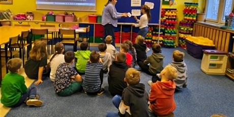 Powiększ grafikę: Grupa dzieci siedzi na dywanie w świetlicy. Na tle granatowej tablicy stoi nauczycielka, któa  podaje dziewczynce kartkę. 