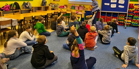 Powiększ grafikę: Grupa dzieci siedzi na dywanie w świetlicy. Trzy osoby mają rękę uniesioną do góry. Z tyłu stoi nauczycielka, obok jest granatowa tablica z informacjami na temat legend.