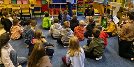 Powiększ grafikę: Grupa dzieci siedzi na dywanie w świetlicy. Z tyłu stoi nauczycielka, która na granatowej tablicy wiesza informacje na lemat legend.