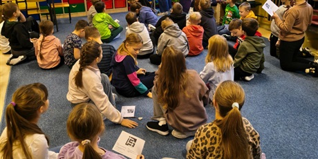 Powiększ grafikę: Grupa dzieci siedzi na dywanie w świetlicy. Z tyłu stoi nauczycielka, która na granatowej tablicy wiesza informacje na lemat legend.