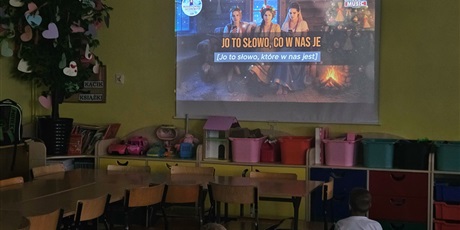 Powiększ grafikę: Grupa dzieci siedzi na dywanie tyłem do obiektywu. Wszyscy wpatrują się w ścienny ekran z prezentacją o muzyce kociewskiej.