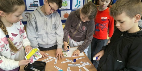 Powiększ grafikę: Grupa dzieci stoi przy stoliku i przykleja na kartki kolorowanki oraz napisy w języku kociewskim. Za nimi znajduje się granatowa tablica z kolorowymi obrazkami podpisanymi w języku kociewskim.