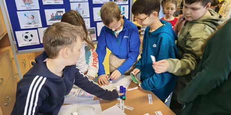 Powiększ grafikę: Grupa dzieci stoi przy stoliku i przykleja na kartki kolorowanki oraz napisy w języku kociewskim. Za nimi znajduje się granatowa tablica z kolorowymi obrazkami podpisanymi w języku kociewskim.