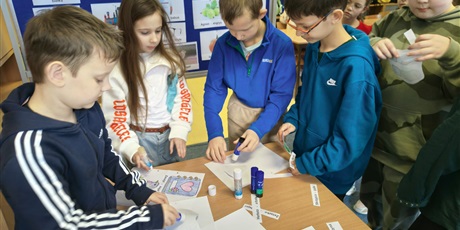 Powiększ grafikę: Grupa dzieci stoi przy stoliku i przykleja na kartki kolorowanki oraz napisy w języku kociewskim. Za nimi znajduje się granatowa tablica z kolorowymi obrazkami podpisanymi w języku kociewskim.