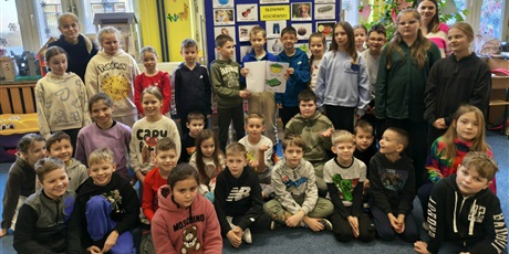 Powiększ grafikę: Grupa dzieci wraz z nauczycielkami pozuje do zdjęcia. Dwóch chłopców trzyma SŁWNIK KOCIEWSKI. Za nimi znajduje się granatowa tablica z kolorowymi obrazkami podpisanymi w języku kociewskim.