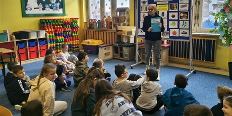 Powiększ grafikę: Nauczycielka stoi obok granatowej tablicy z kolorowymi obrazkami podpisanymi w języku kociewskim. W ręku trzyma słownik kociewski. Grupa dzieci siedzi na dywanie zwrócona w stronę nauczycielki.