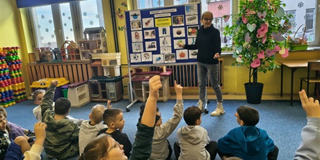 Powiększ grafikę: Nauczycielka stoi obok granatowej tablicy z kolorowymi obrazkami podpisanymi w języku kociewskim. W ręku trzyma słownik kociewski. Grupa dzieci siedzi na dywanie zwrócona w stronę nauczycielki.