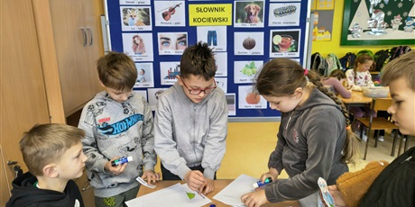 Powiększ grafikę: Grupa dzieci stoi przy stoliku i przykleja na kartki kolorowanki oraz napisy w języku kociewskim. Za nimi znajduje się granatowa tablica z kolorowymi obrazkami podpisanymi w języku kociewskim.