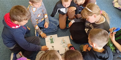 Powiększ grafikę: grupa dzieci w kole pochyla się nad plakatami ze strojami kaszubskimi. Przyklejają hasła z elementami strojów.