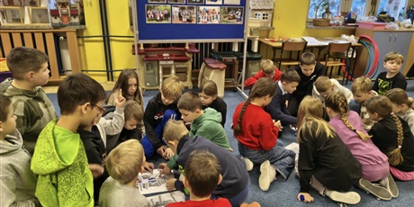 Powiększ grafikę: Dwie grupy dzieci siedzą w dwóch kręgach na dywanie. Dzieci pochylają się nad plakatami, które leżą między nimi. Za nimi stoi granatowa tablica informacyjna dotycząca strojów kociewskich.