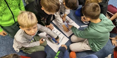 Powiększ grafikę: Dzieci pochylają się nad plakatami, które leżą między nimi. Uczniowie przyklejają na nie karteczki z nazwami poszczególnych elementów strojów kociewskich.